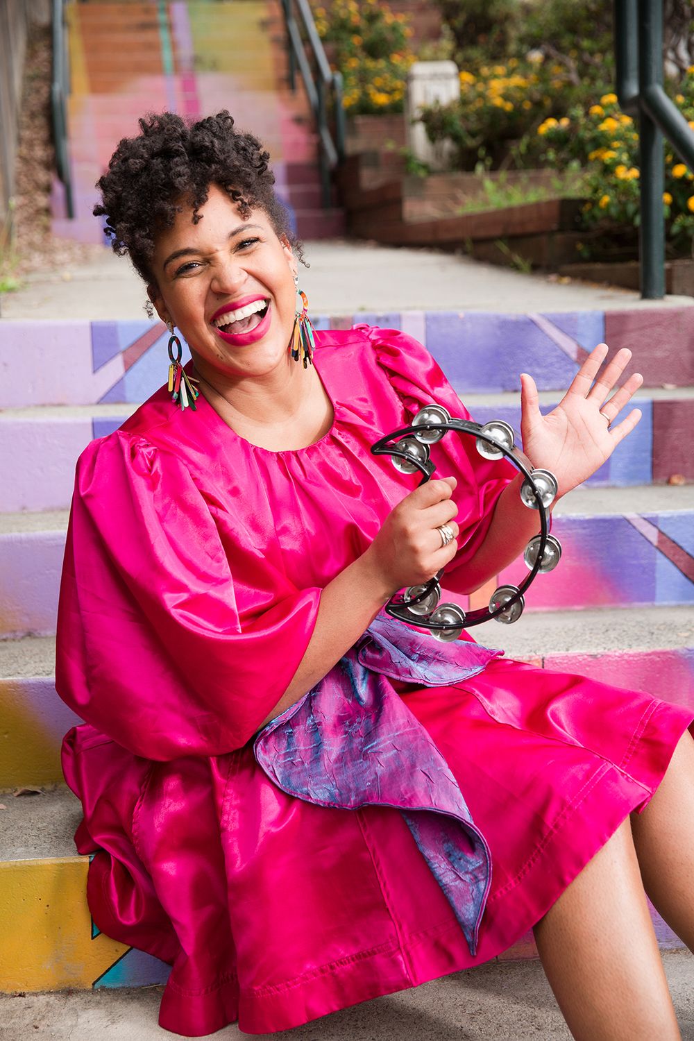 A joyful woman in a vibrant pink dress sits on colorful steps, holding a tambourine and waving.