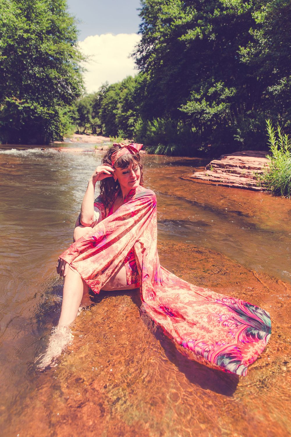 Woman in pink paisley wrap sitting on rocks in a shallow river surrounded by lush green trees.