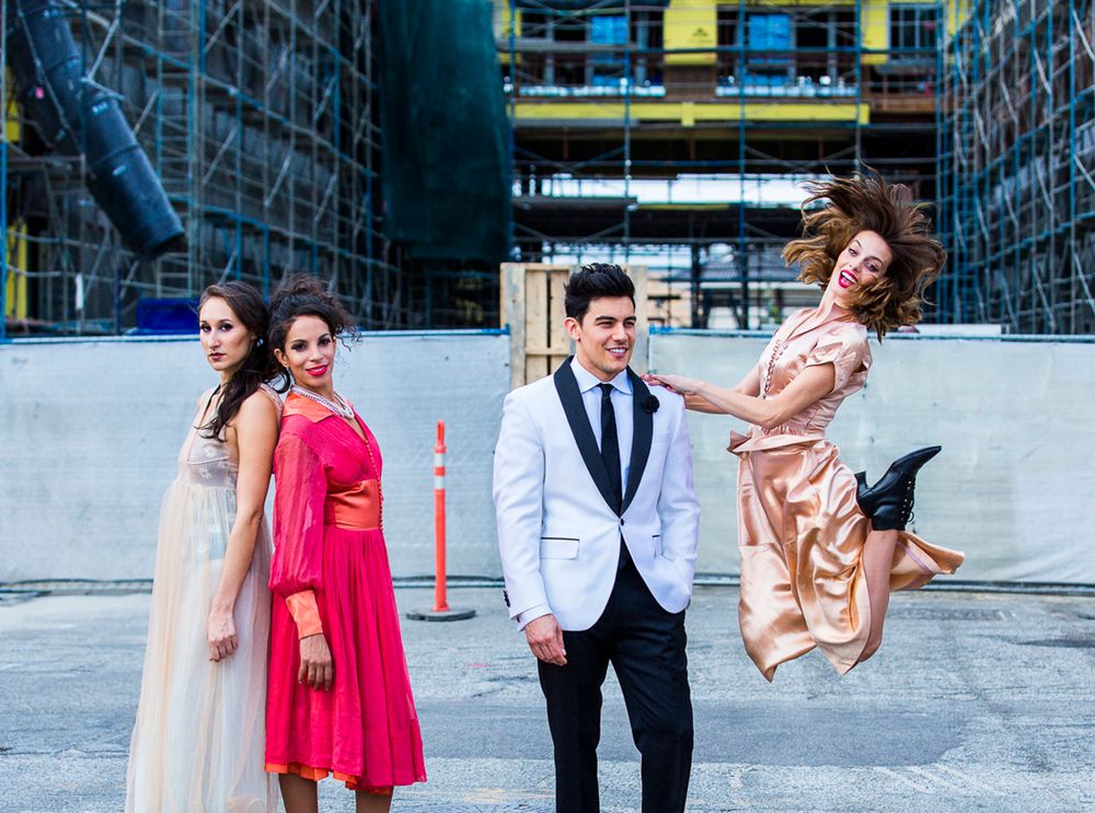 Four elegantly dressed people pose at a construction site, with one woman joyfully jumping in a gold dress.