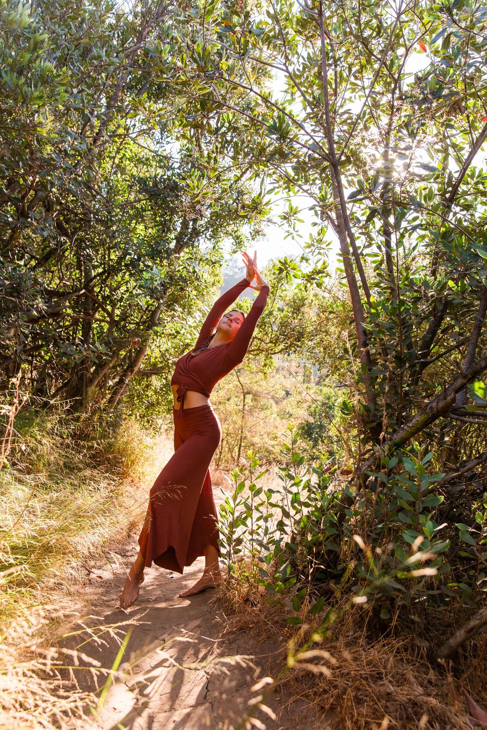 Woman in red dress dancing on a sunlit forest trail surrounded by lush green trees.