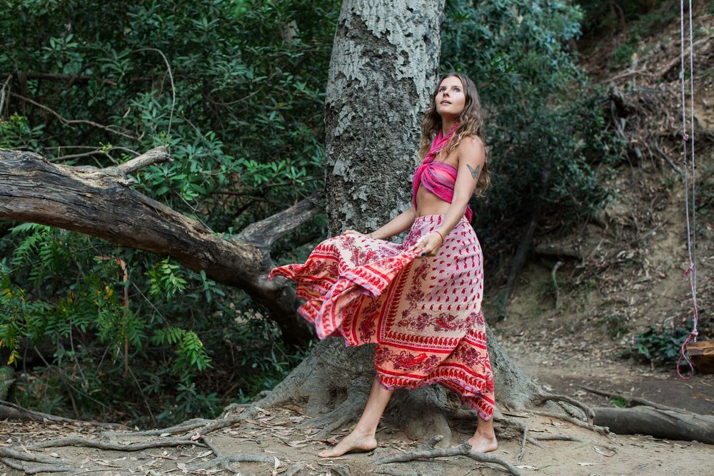 Woman in red floral maxi skirt and halter top poses barefoot among trees and roots in a forest setting.