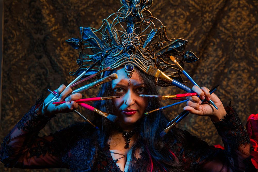 Woman in elaborate fantasy headdress holding crossed arrows, dramatic costume with blue lighting.