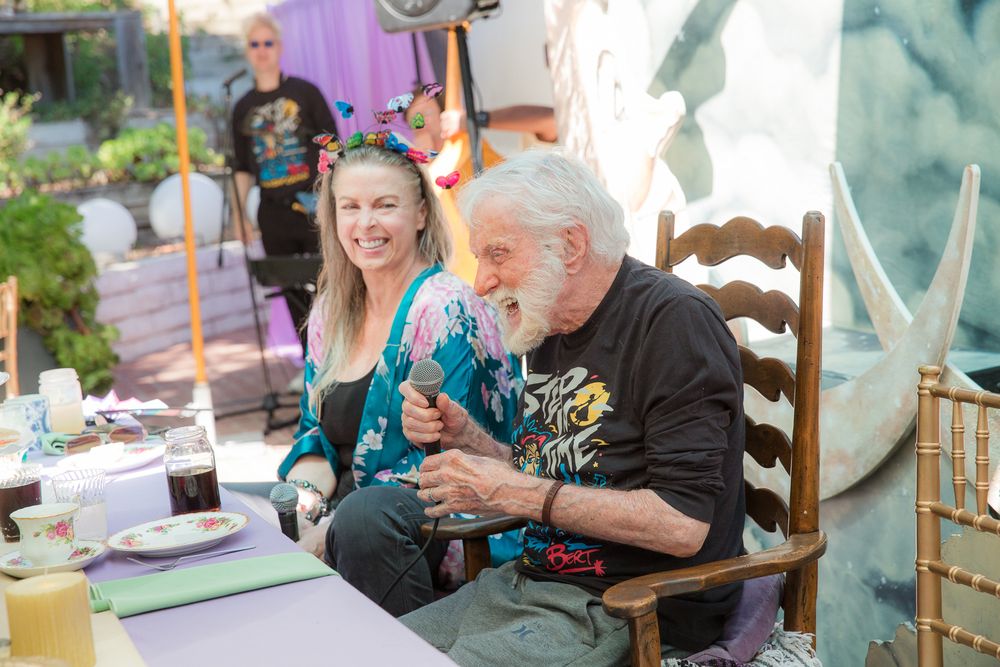 Two people smiling and laughing together at an outdoor table with colorful decorations in the background.