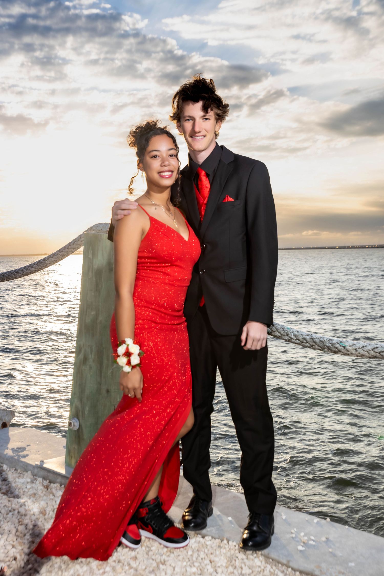 A couple dressed in prom attire poses by the water at sunset, wearing red dress and black tuxedo.