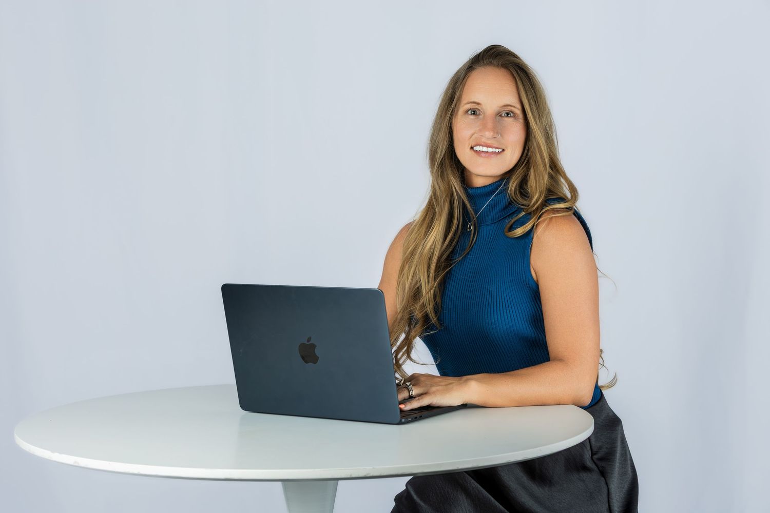 Smiling woman in blue turtleneck top sitting at white table with MacBook laptop against light gray background.