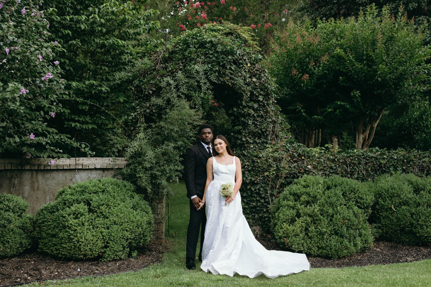 A bride and groom pose together in a lush garden setting, with the bride holding a bouquet in her white wedding gown.