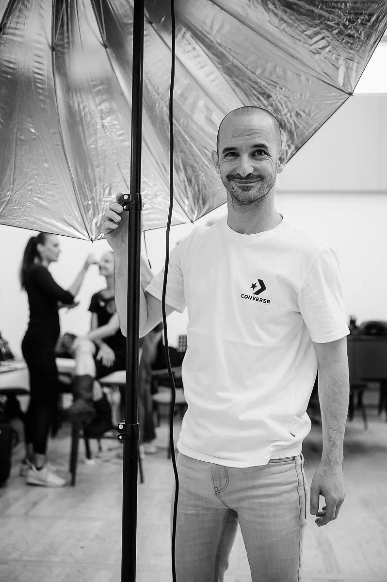 Smiling bald man in white Converse t-shirt stands by photography umbrella light in black and white studio photo.