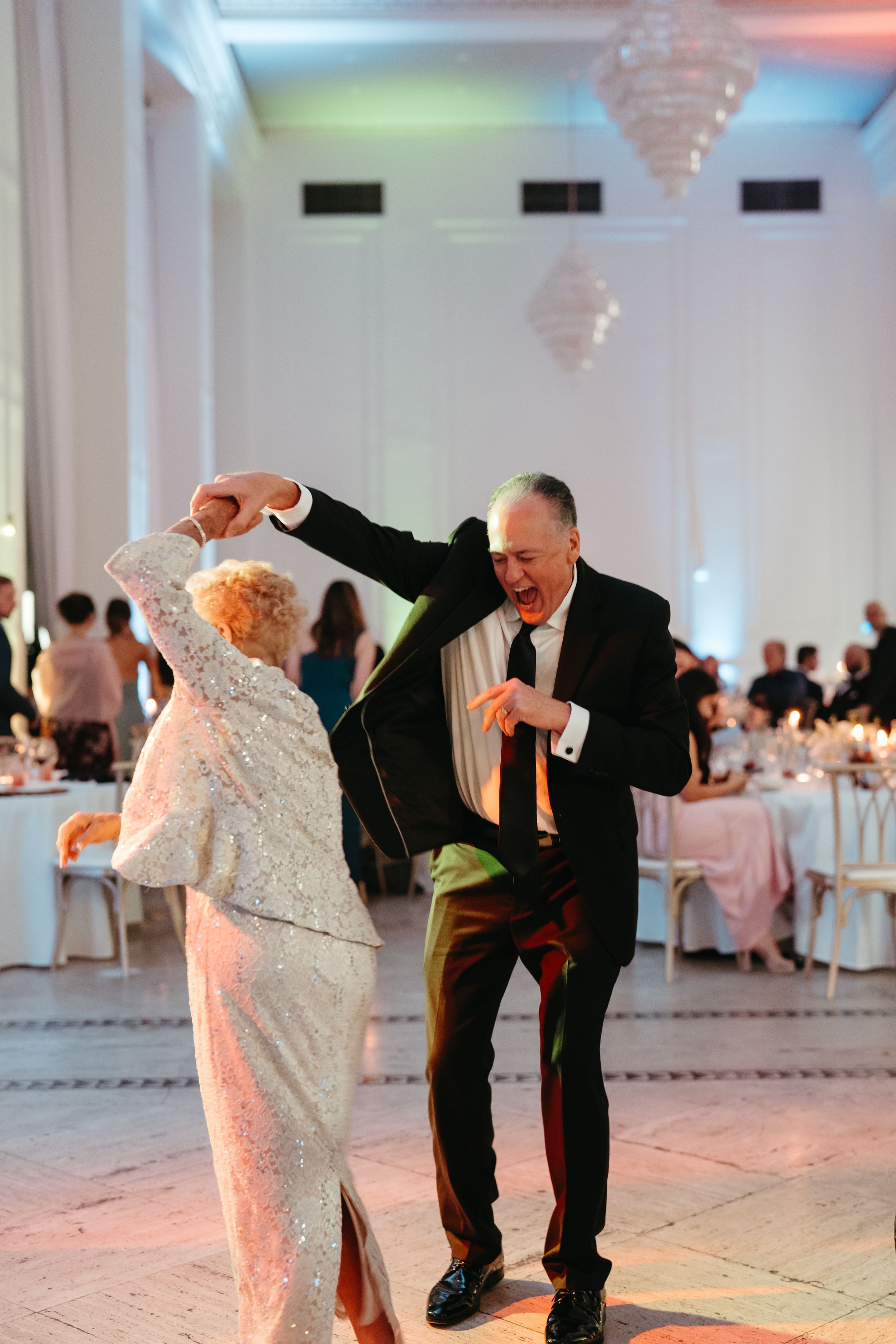 Elderly couple dancing joyfully at a wedding reception in an elegant ballroom with chandeliers and guests seated at tables.