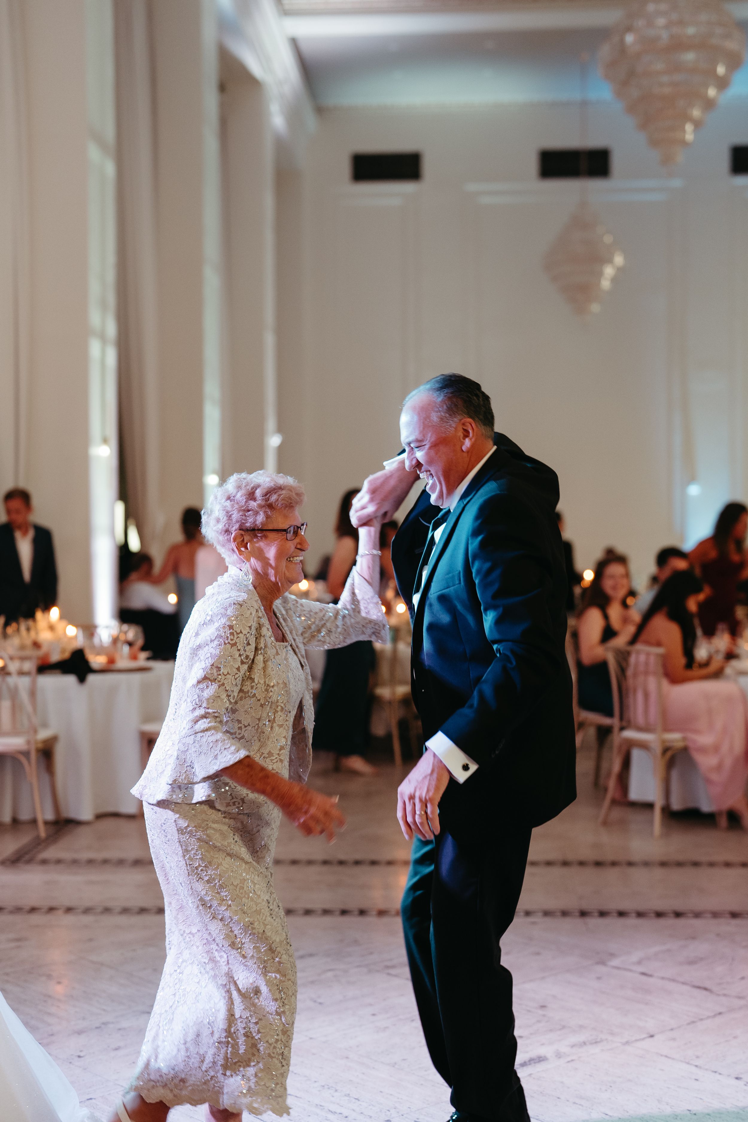 Elderly woman and man in formal attire share a joyful dance at an elegant wedding reception.