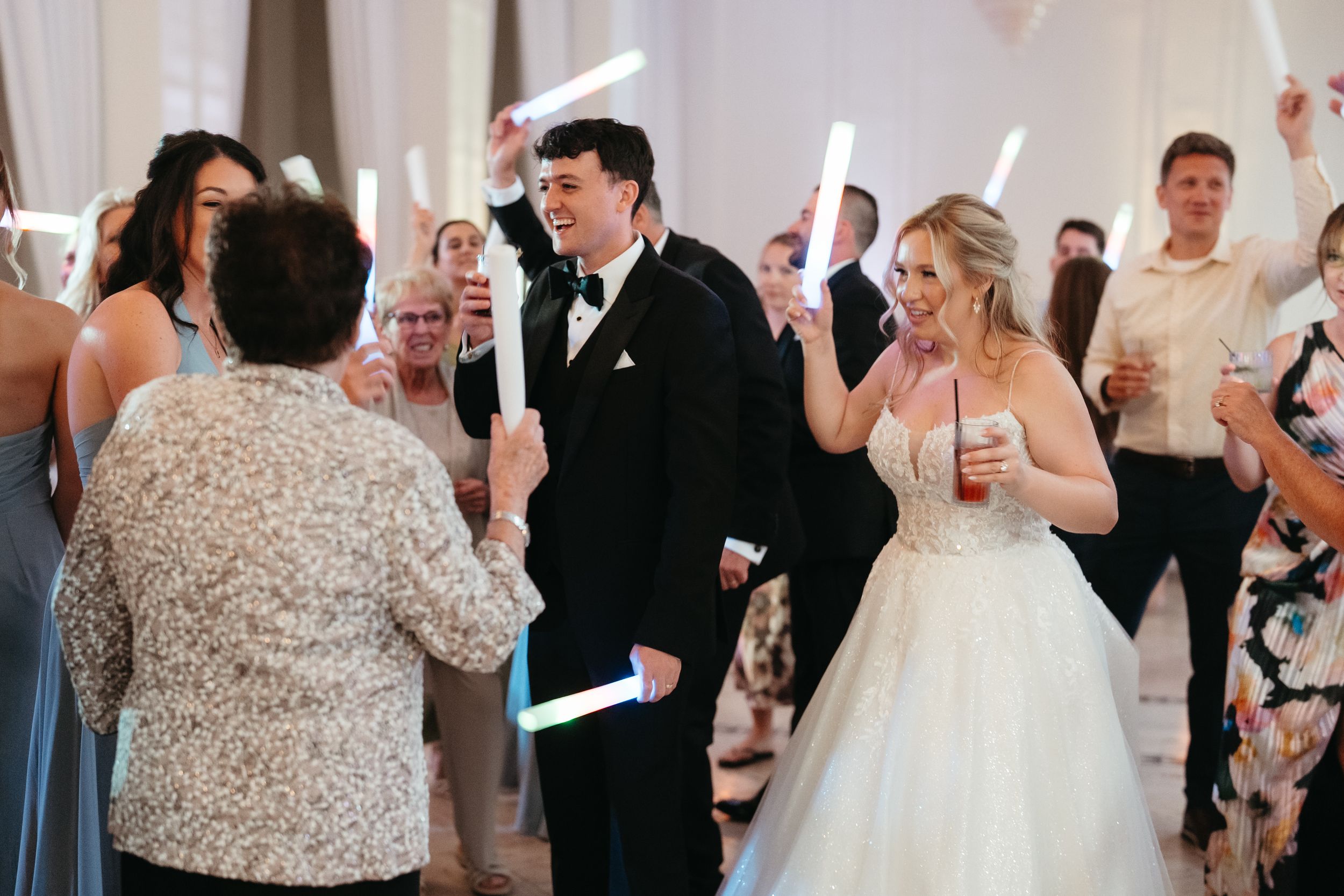Bride and groom dancing at wedding reception surrounded by guests holding glowing light sticks in elegant white venue.