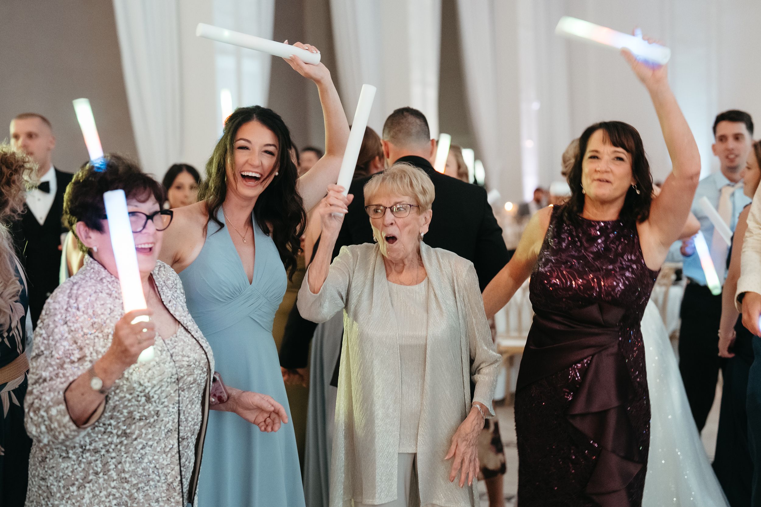 Four women joyfully wave glowing foam sticks at a wedding reception in an elegant white venue.