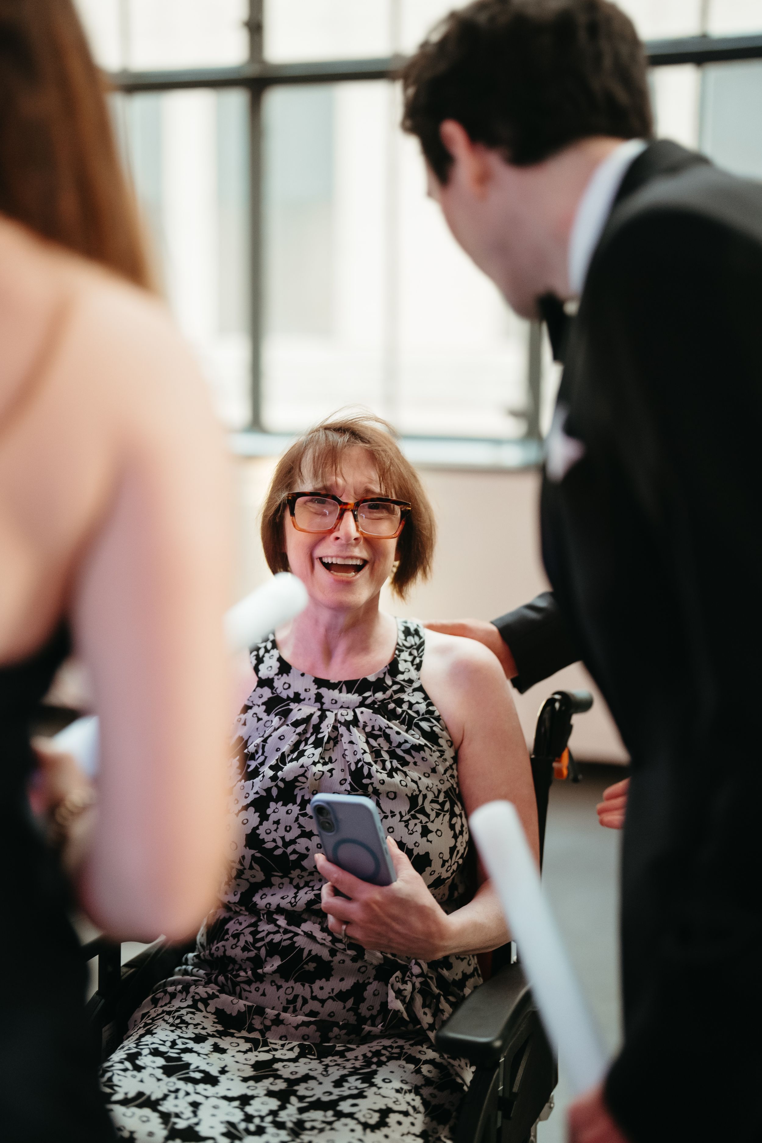 Elderly woman in wheelchair wearing glasses and floral dress, smiling while holding phone at formal event.