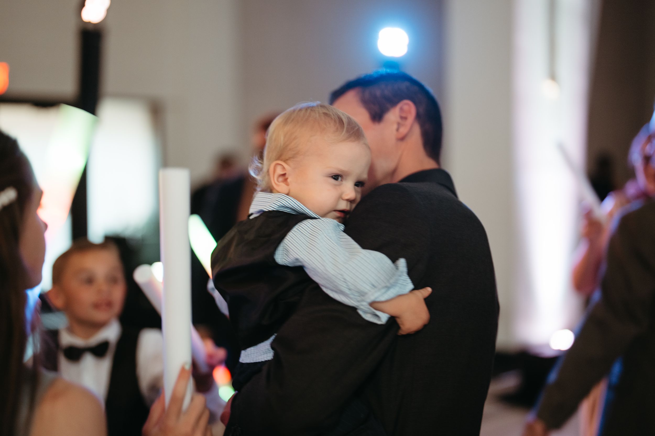 Man in black suit holding a baby dressed in formal attire at an elegant event with candles.