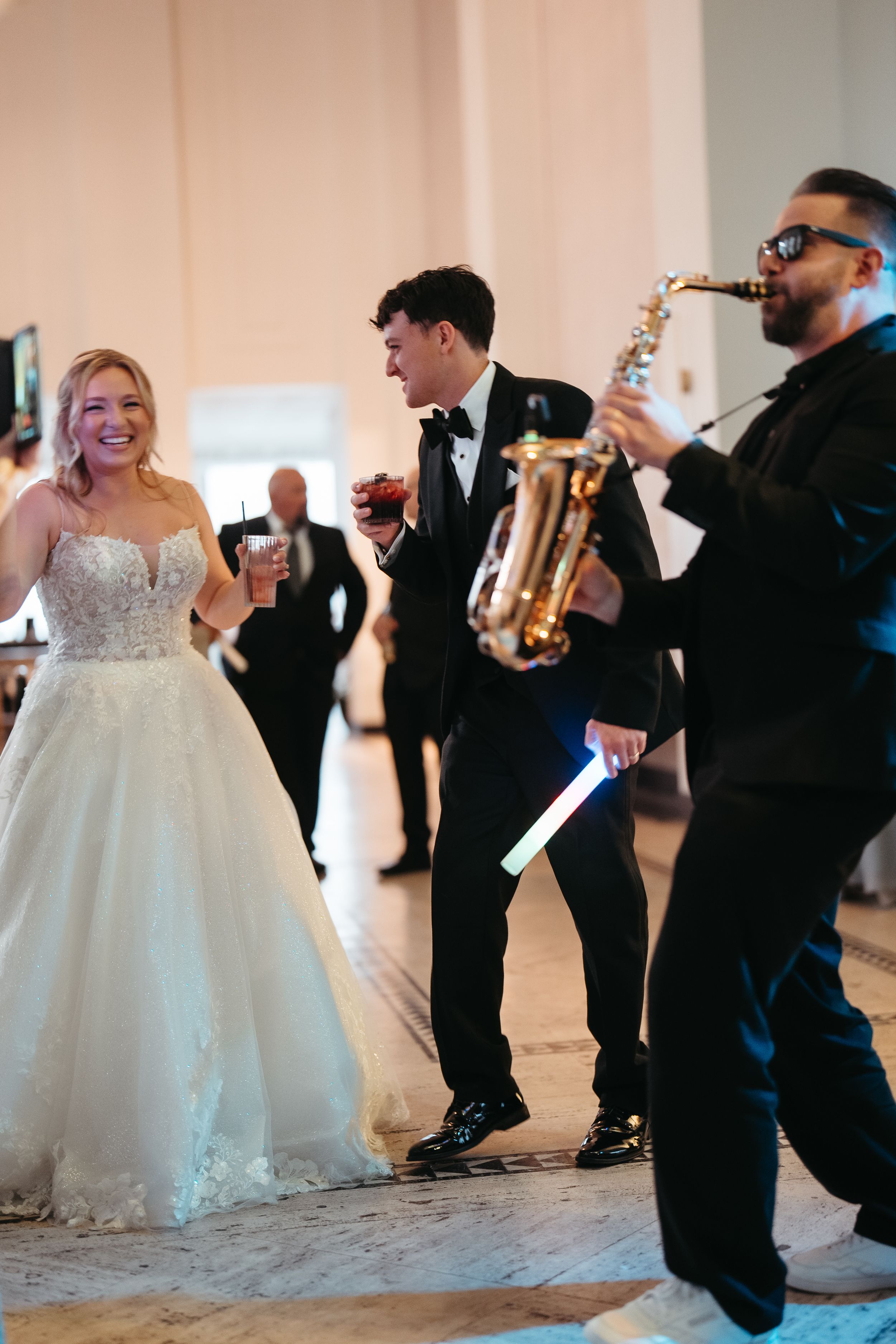Bride in white gown and groom in tuxedo dancing with saxophonist at elegant wedding reception.