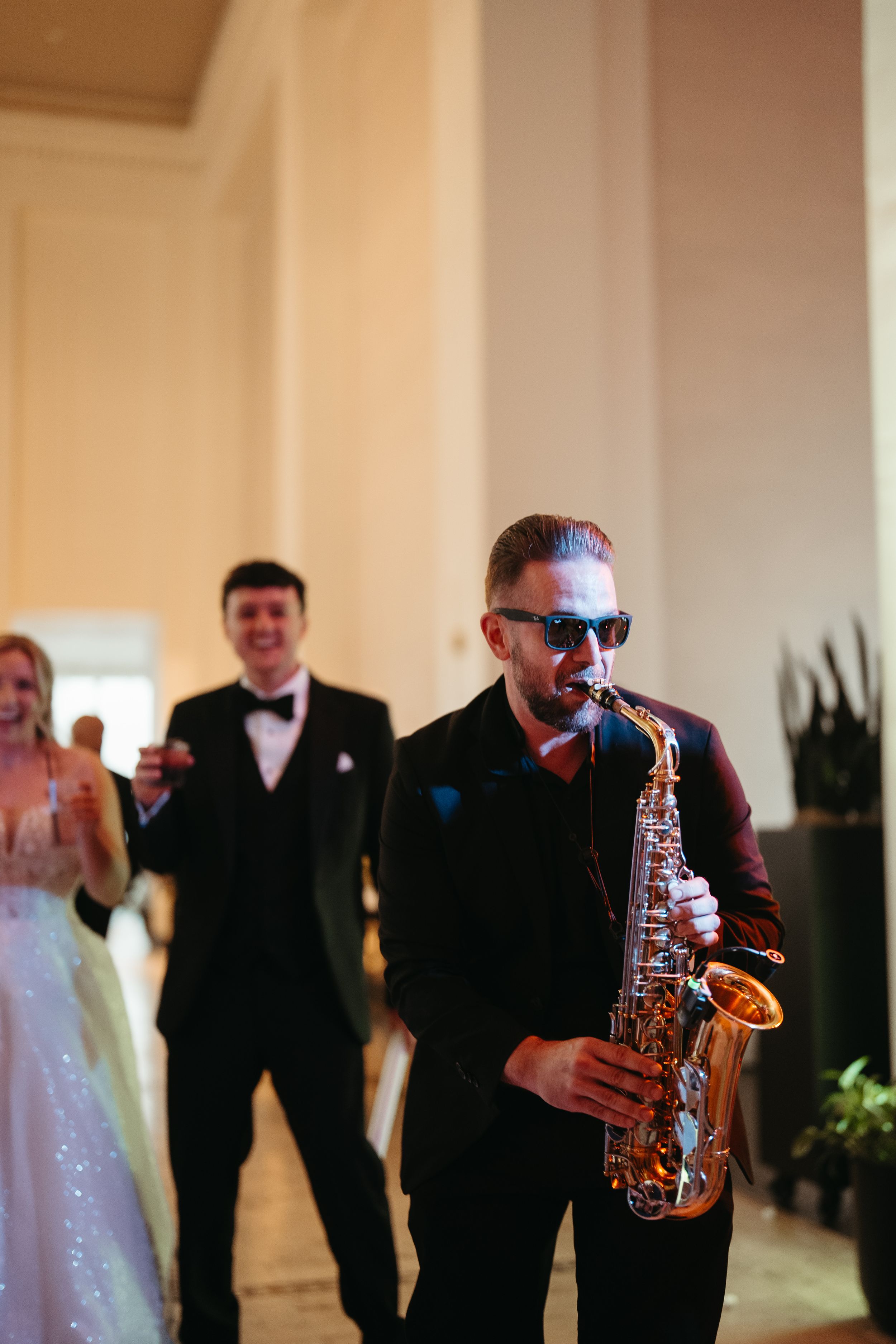 Saxophonist in sunglasses performs at elegant wedding reception while guests celebrate in background.