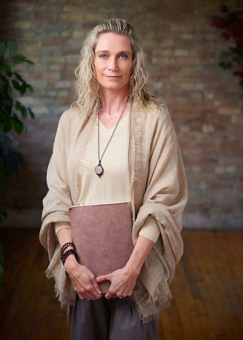 Woman with blonde hair wearing a beige shawl and pendant necklace holds a folder, standing before a brick wall.