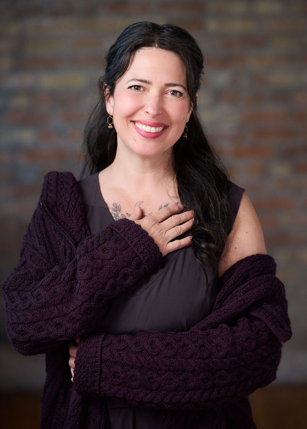 Smiling woman with dark hair wearing a purple knit cardigan and gray dress, posing warmly against a brick wall.