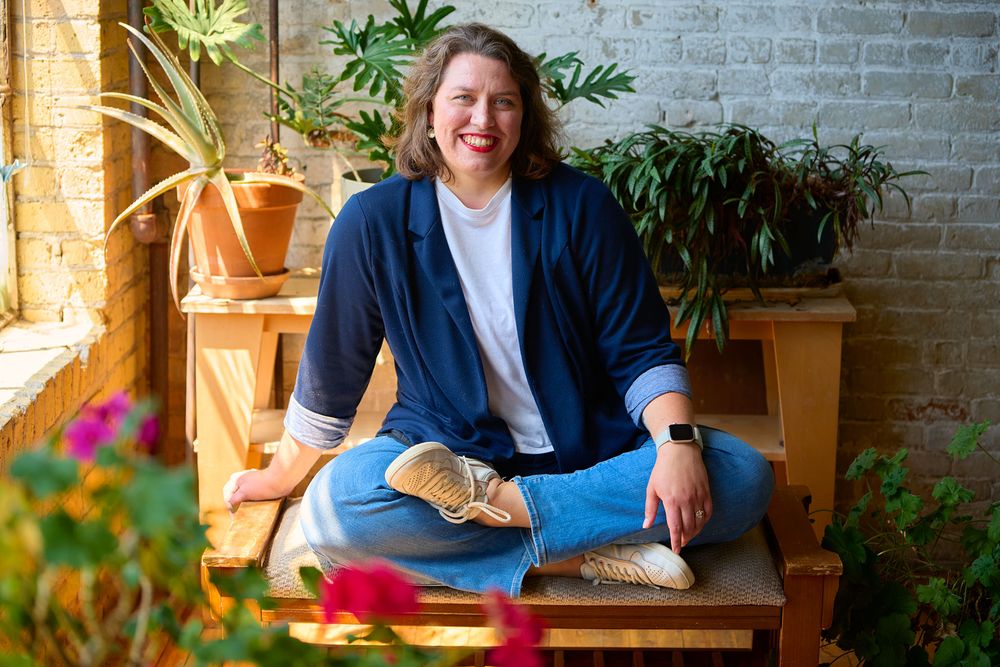 Woman in blue blazer and jeans sits cross-legged on wooden bench surrounded by lush indoor plants.