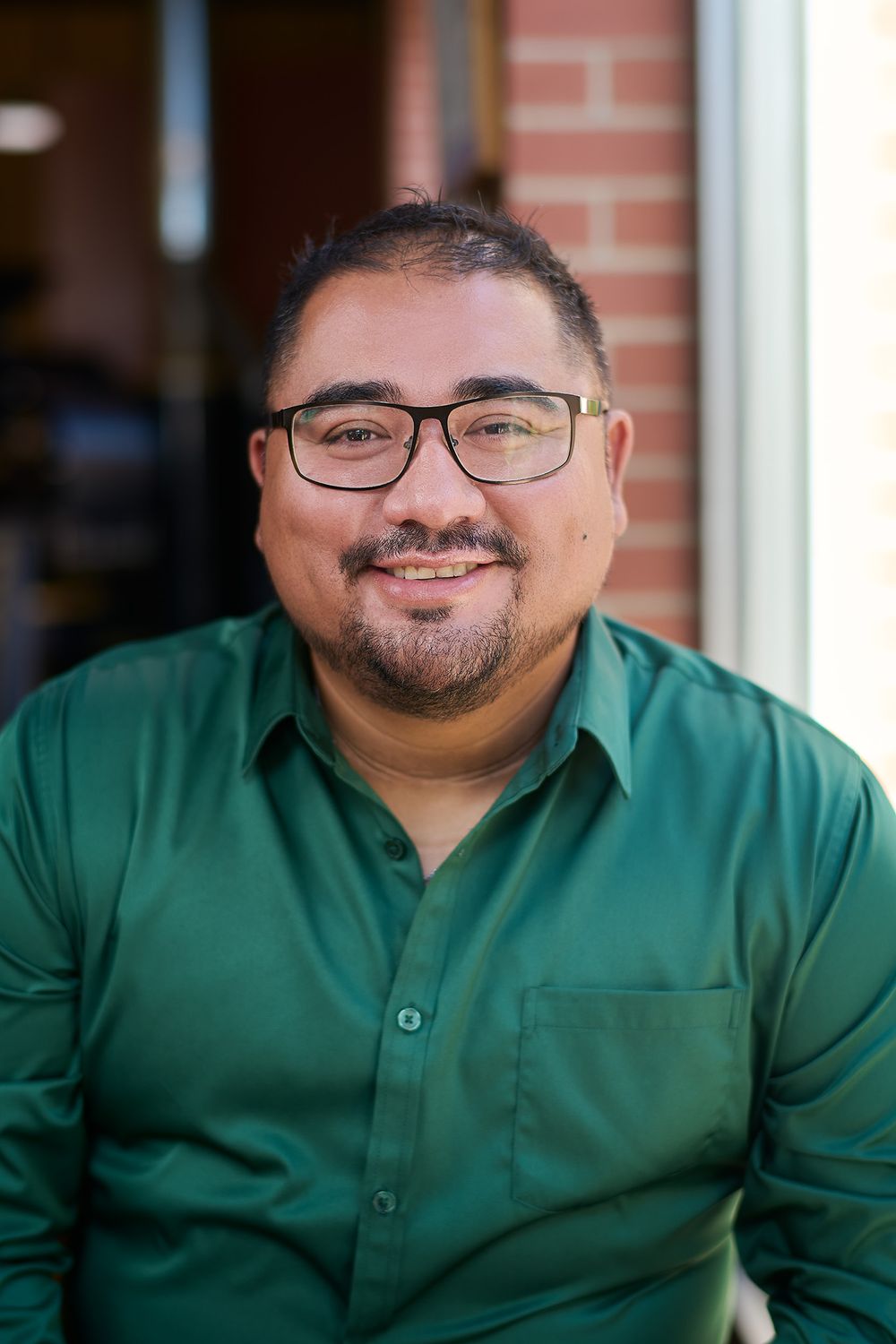 Smiling man with glasses and green button-up shirt poses outdoors near brick wall.