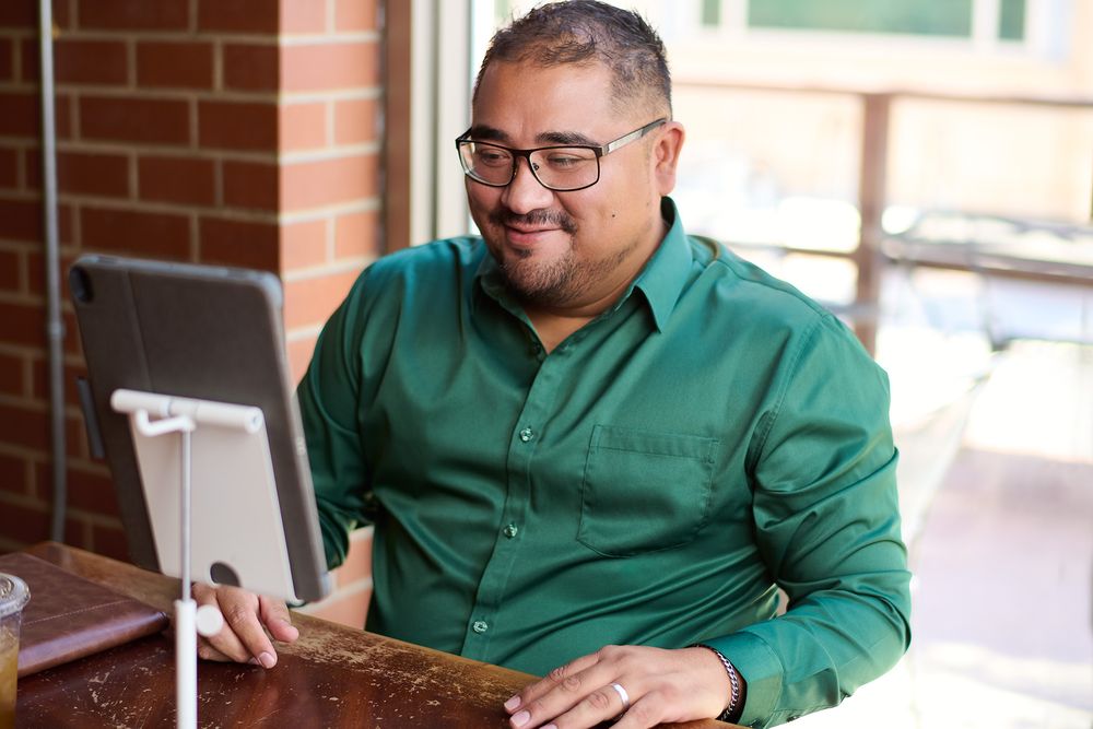 Man in green shirt smiling at tablet on stand, sitting at wooden desk in bright office space.