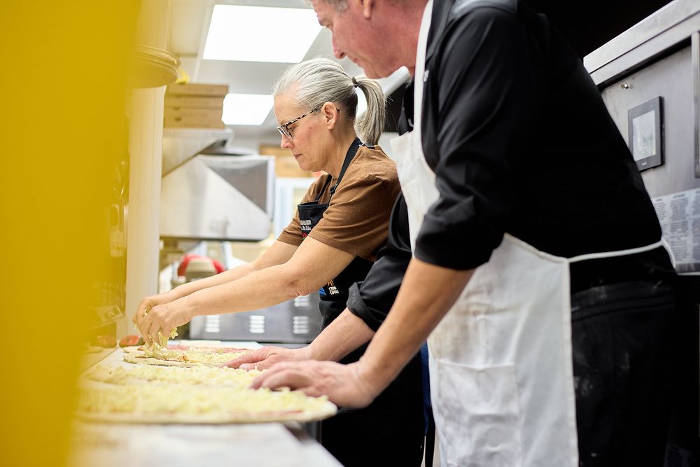 Two chefs in black aprons preparing pizza in a professional kitchen, adding toppings to dough.