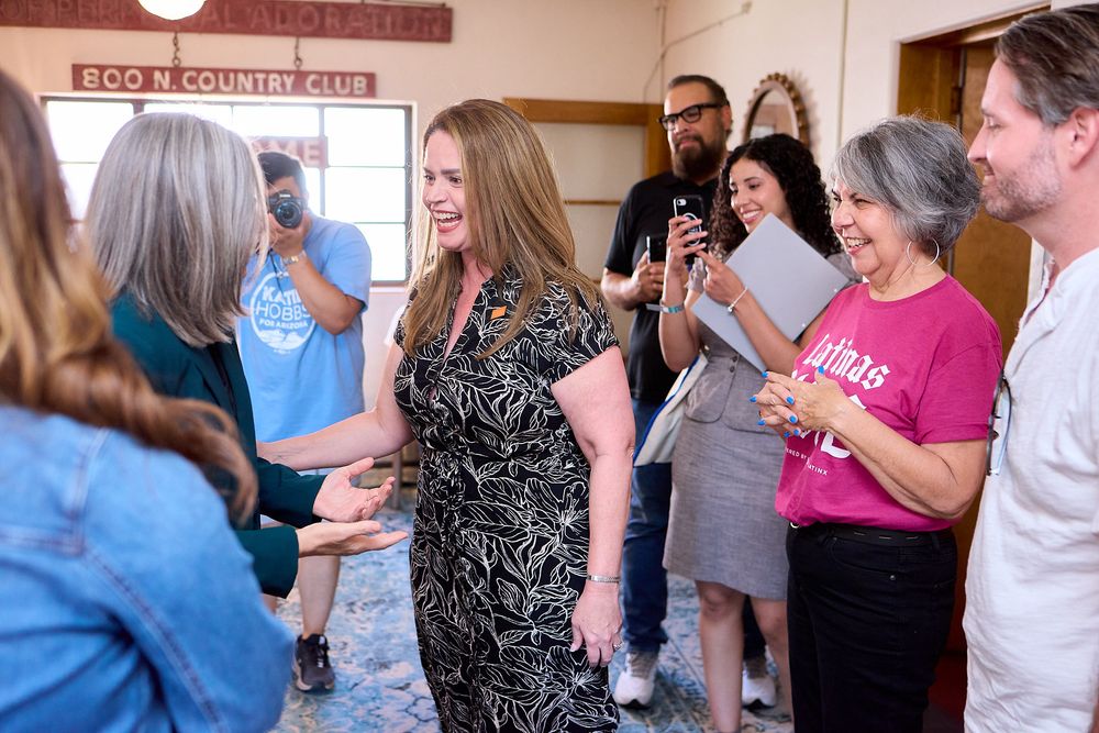 A woman in a floral dress laughs while greeting a crowd at an indoor community gathering event.