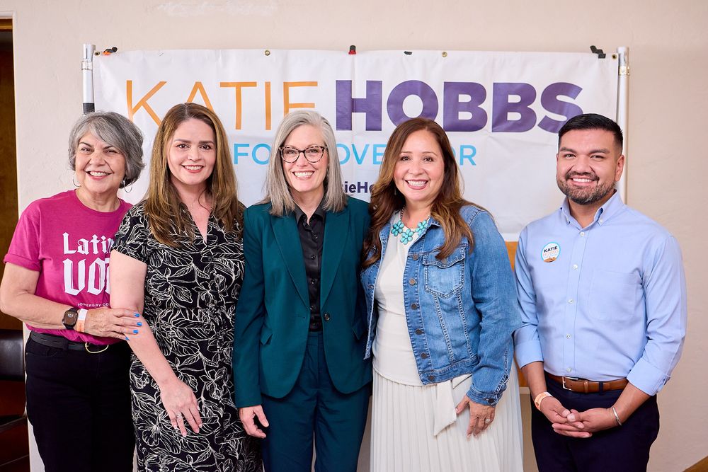 Five people smile together in front of a Katie Hobbs for Governor campaign banner at a political event.