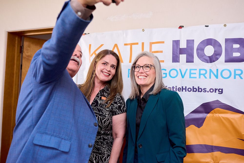 Three people posing for a selfie in front of a Katie Hobbs for Governor campaign banner at a political event.