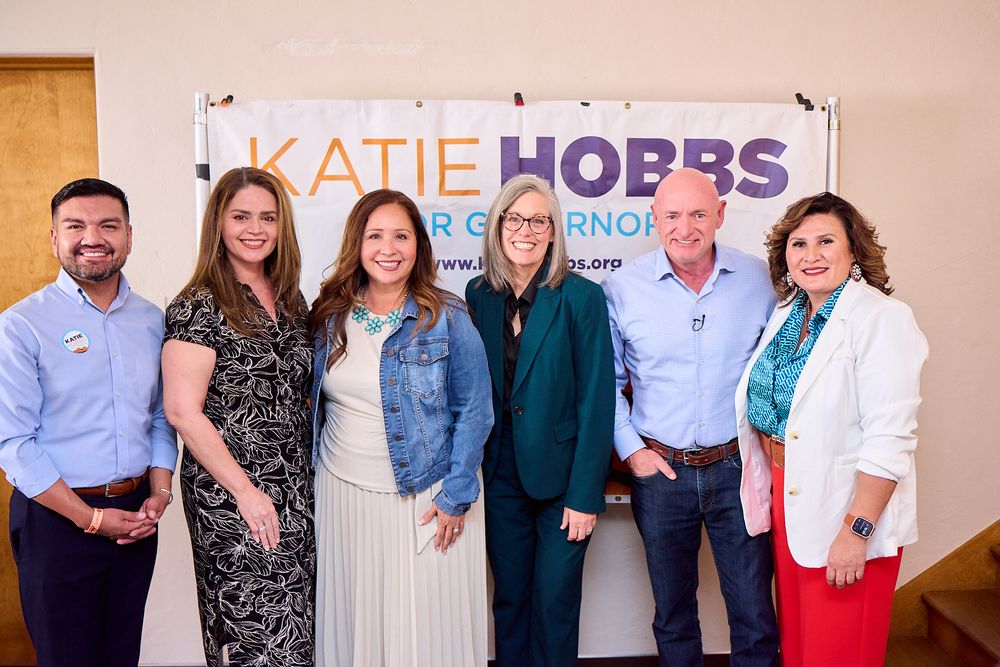 Six people smile together in front of a Katie Hobbs for Governor campaign banner at a political event.