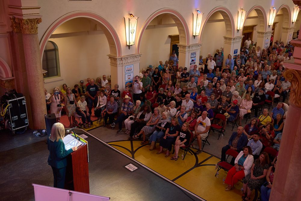 Large crowd gathered in elegant hall listening to female speaker at podium during formal event.