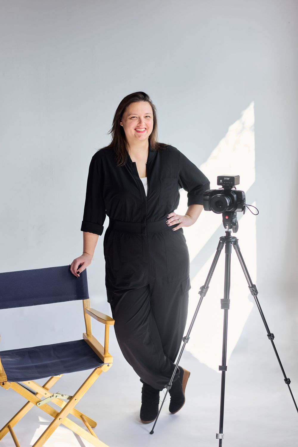 Woman in black outfit smiling beside a director's chair and camera on tripod in a bright white studio.