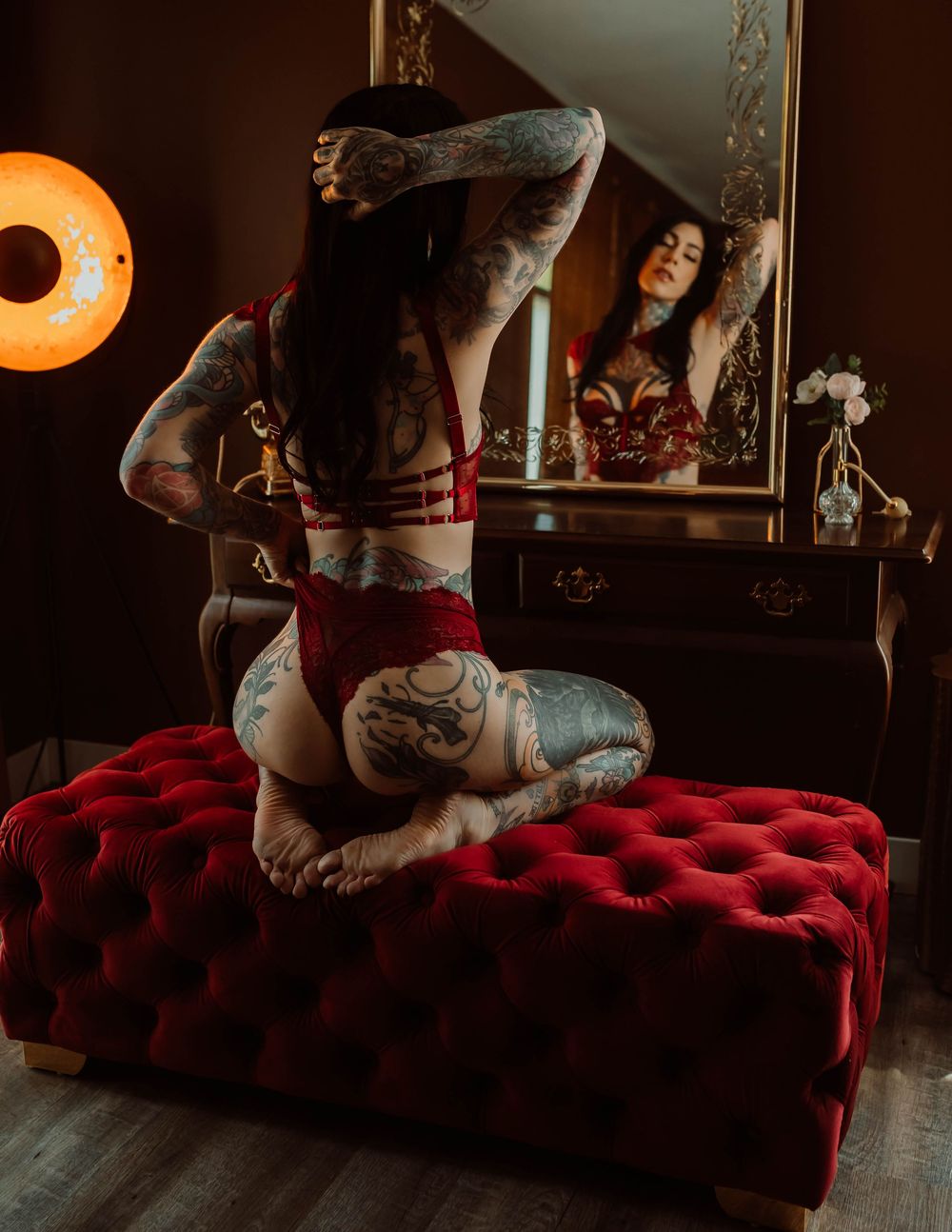 Tattooed woman in red lingerie kneeling on tufted velvet ottoman before vanity mirror in moody boudoir setting.