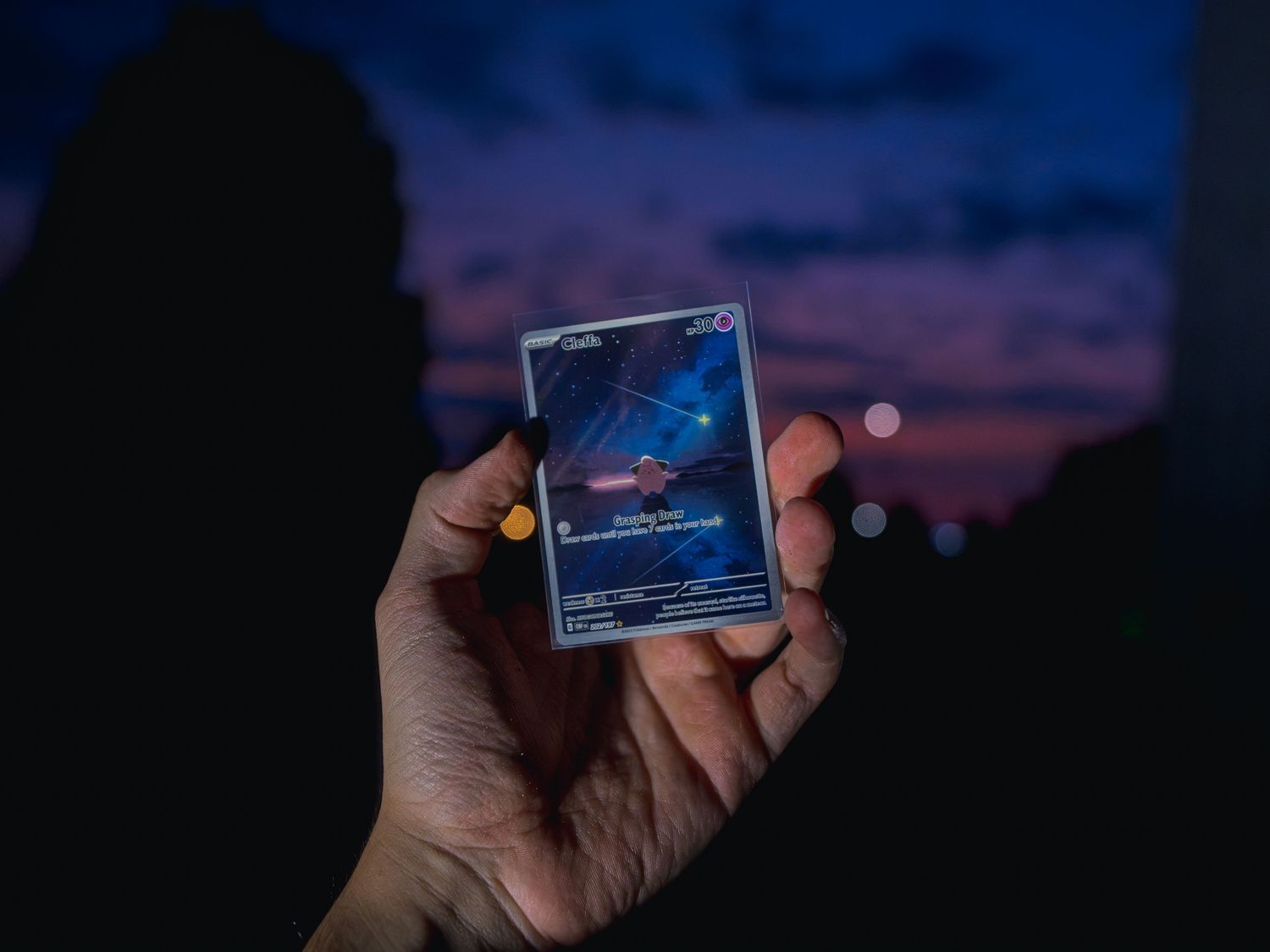 A hand holds a sleeved collectible trading card with starry night-sky artwork against a purple and orange twilight sky with bokeh lights