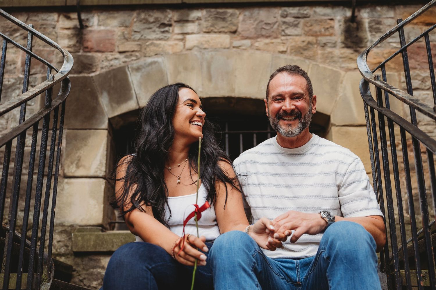 Happy couple smiling outdoors in front of historic buildings, wearing casual sweaters on a cloudy day.