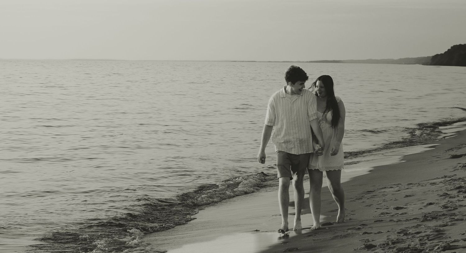 Couple walking hand in hand along a serene beach shoreline in black and white photo.