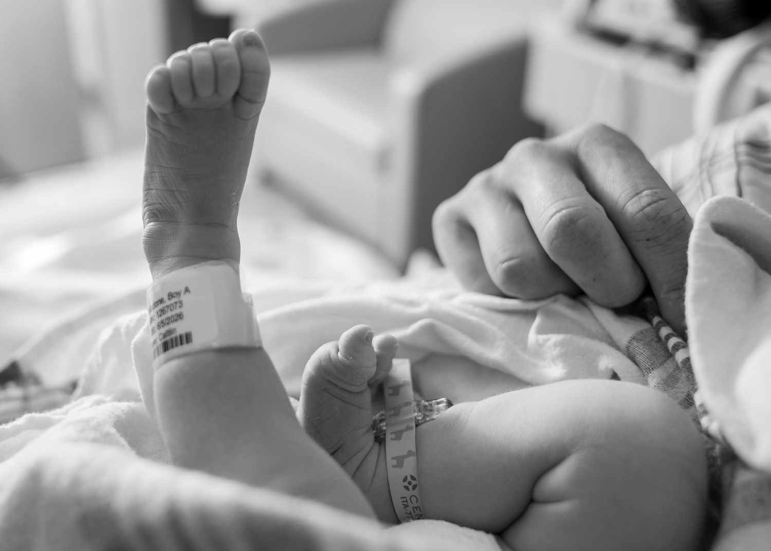 Black and white photo of a newborn baby with hospital ID bracelet, held by parent's hand.