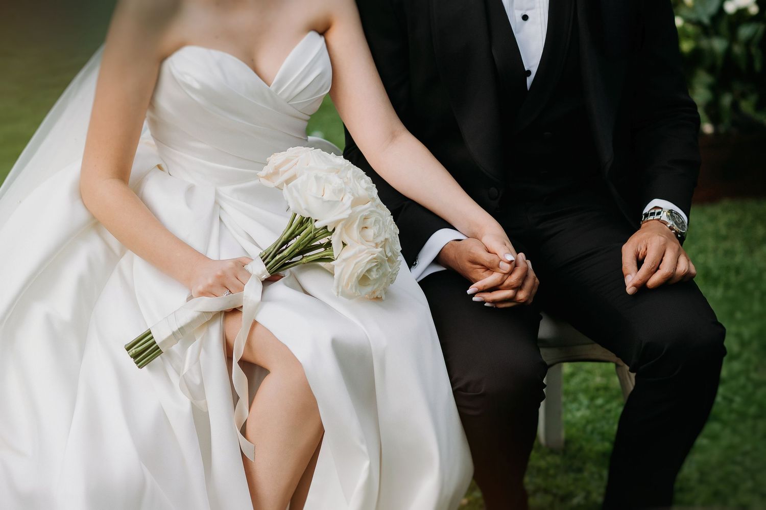 Bride in strapless gown holding white bouquet seated beside groom in tuxedo holding hands outdoors.