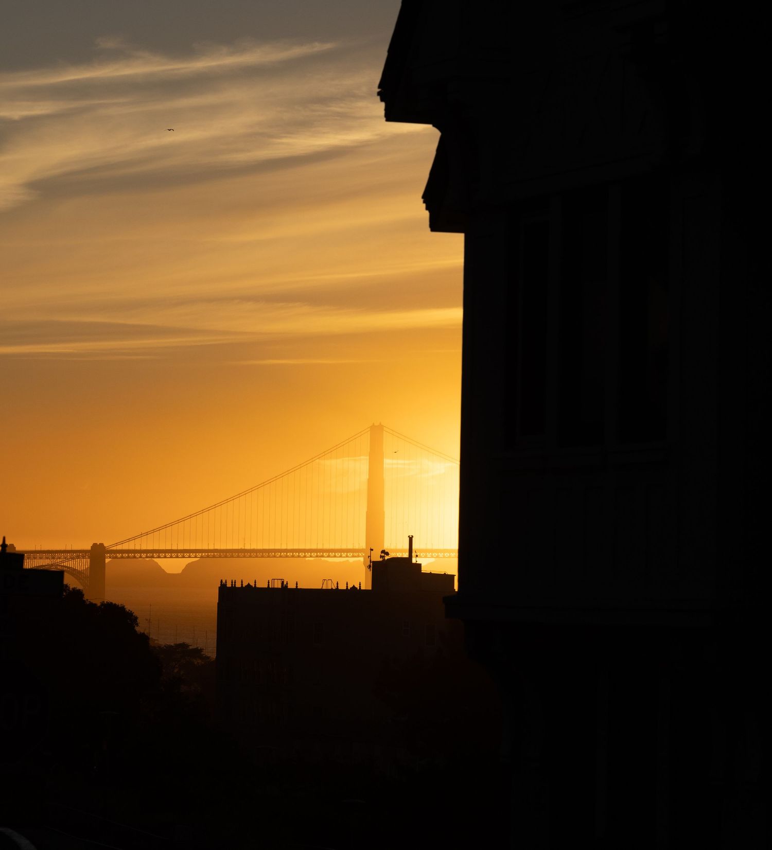 Silhouette of Golden Gate Bridge at sunset with warm orange sky and building foreground in San Francisco.