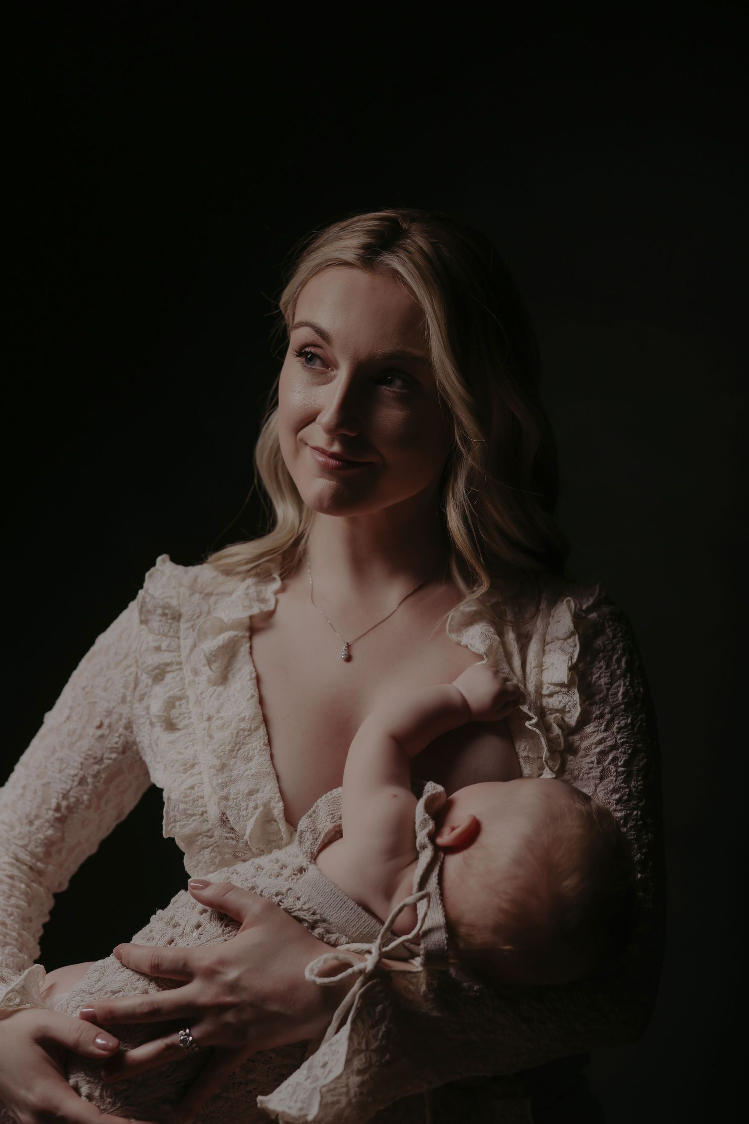 A blonde woman in white lace dress tenderly breastfeeds her newborn baby against a dark studio background.