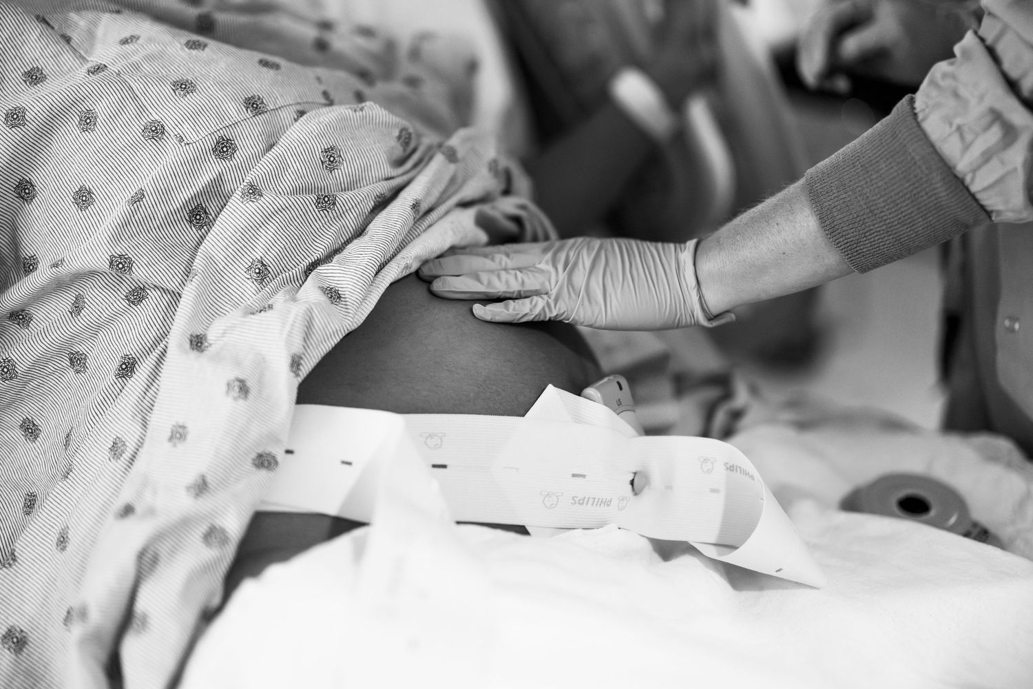 Black and white photo of a woman in a hospital gown leaning on a walker in a medical room.