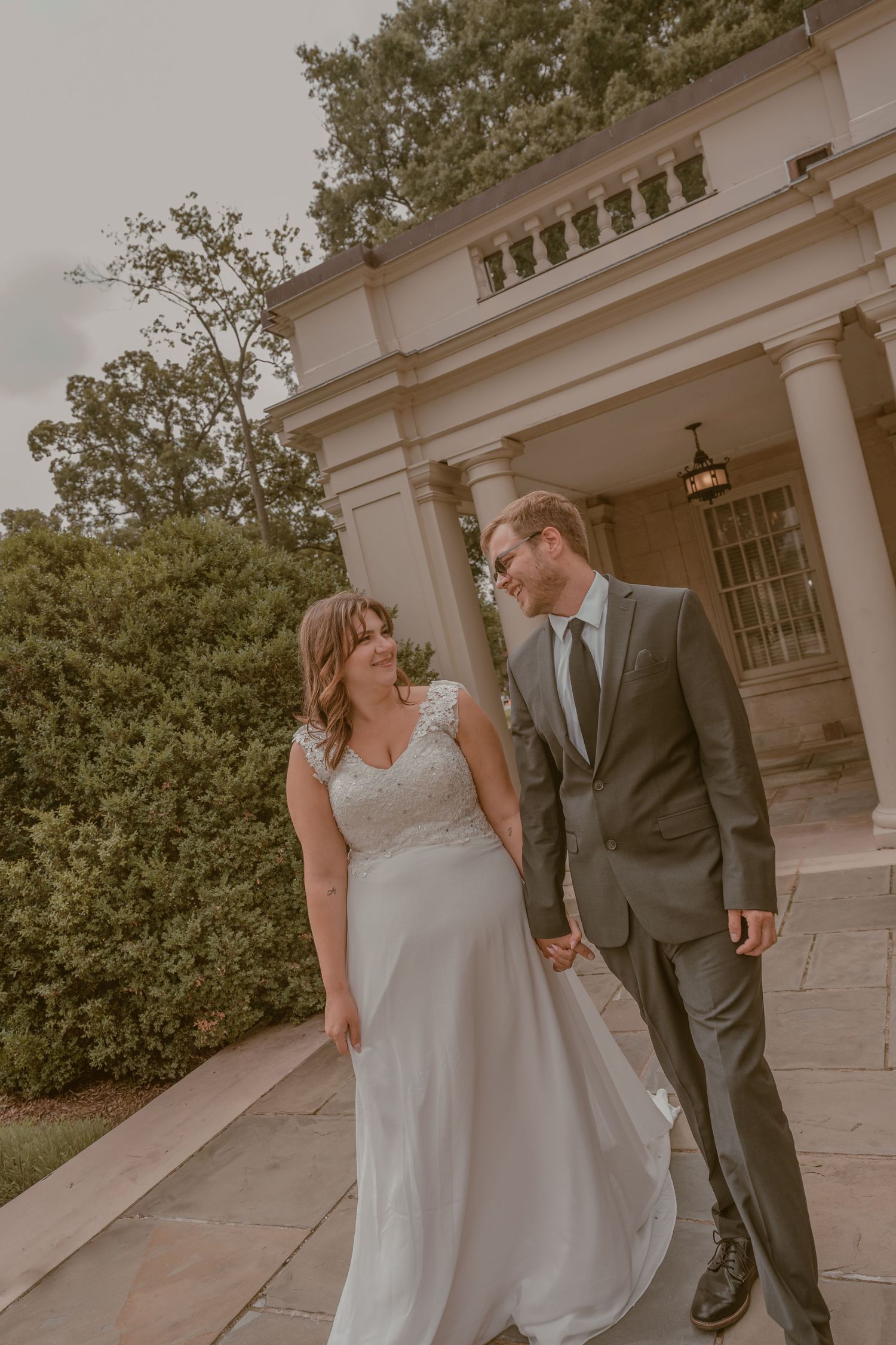Bride and groom walk hand in hand smiling before elegant columned mansion on their wedding day.