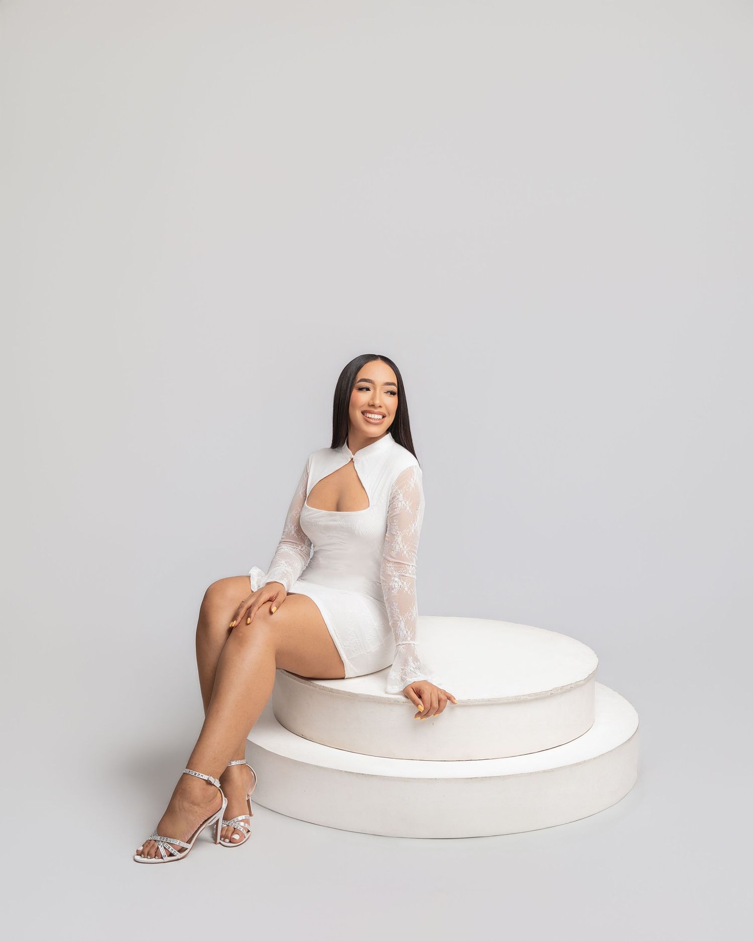 Woman in white cutout mini dress and silver heels sits elegantly on white tiered platform against gray background.