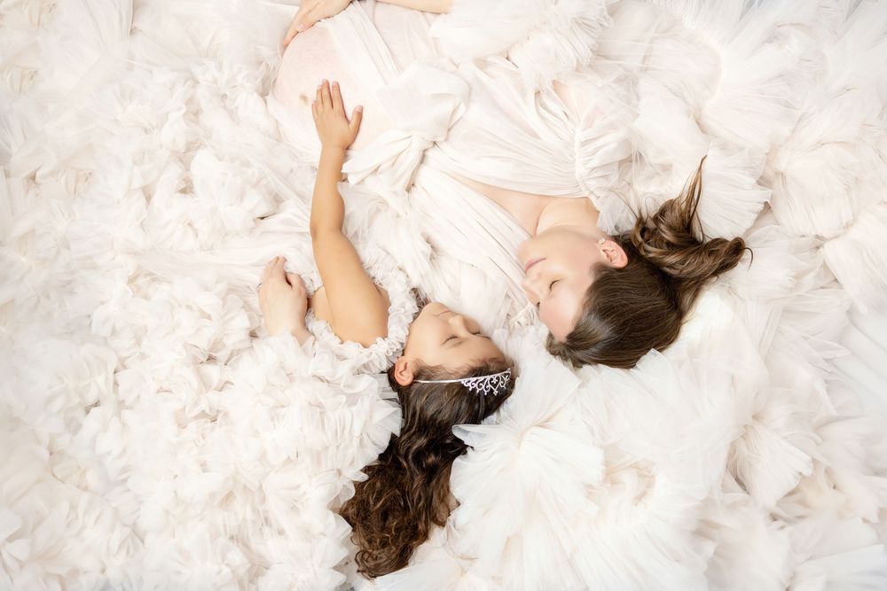 Two girls in white dresses lying on fluffy white feathers, smiling and posing together in a dreamy, ethereal setting.
