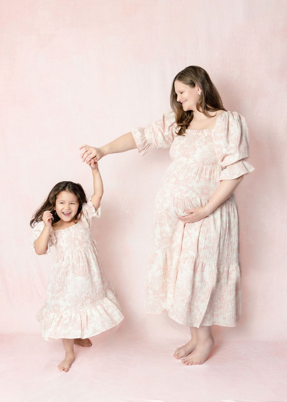 Pregnant woman and young girl in matching pink floral dresses dancing together against a soft pink background.