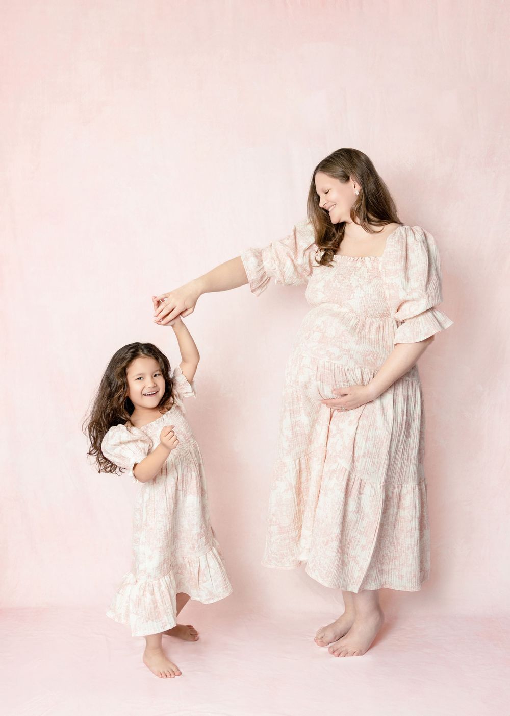 Pregnant mother in floral dress twirling young daughter in matching dress against soft pink background.