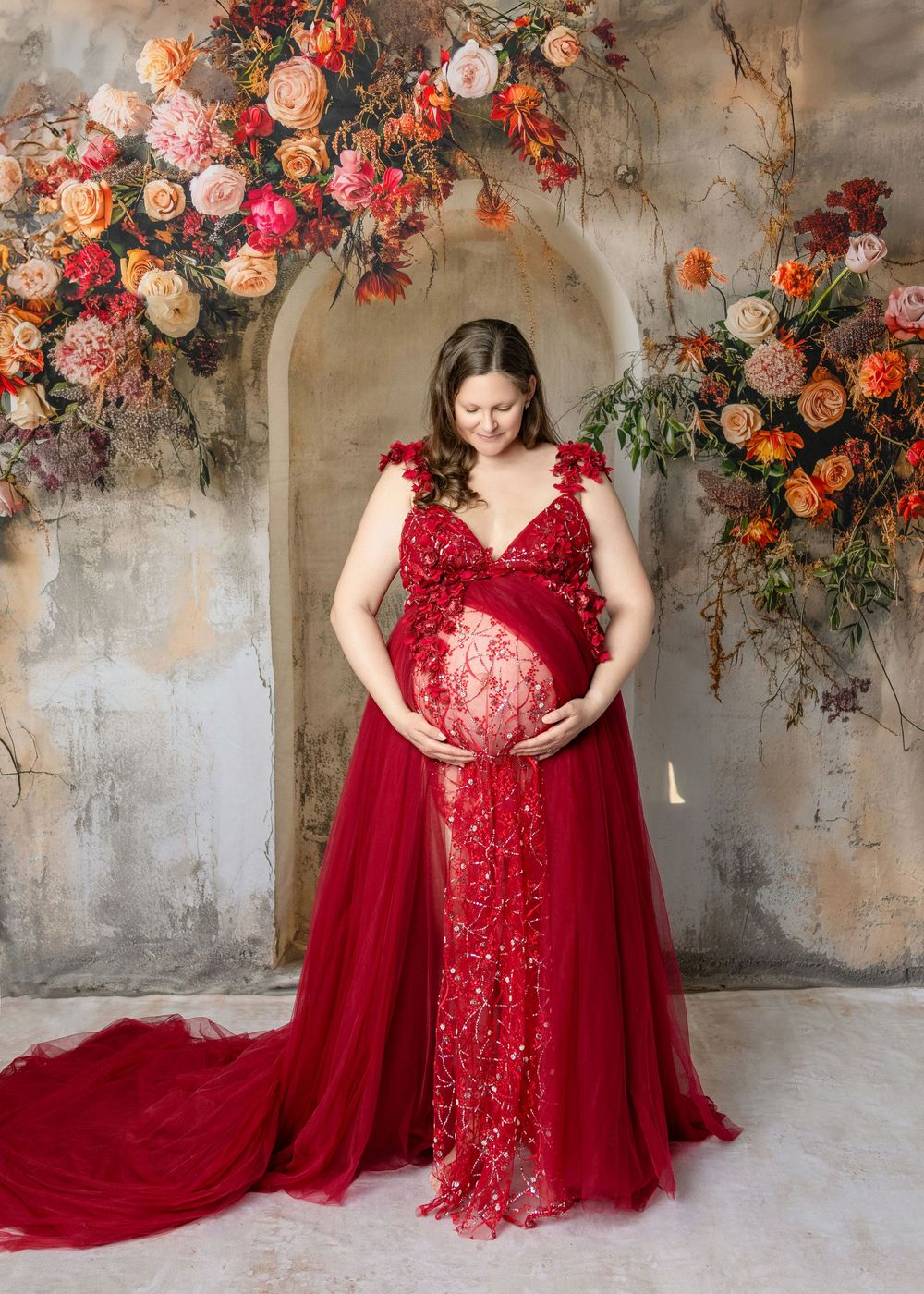 Pregnant woman in red lace gown cradling baby bump, surrounded by vibrant floral arch backdrop in maternity photo.