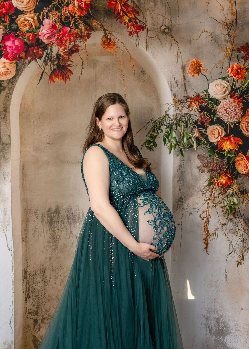 Pregnant woman in teal sequin gown poses before floral arch with orange and pink blooms for maternity photo.