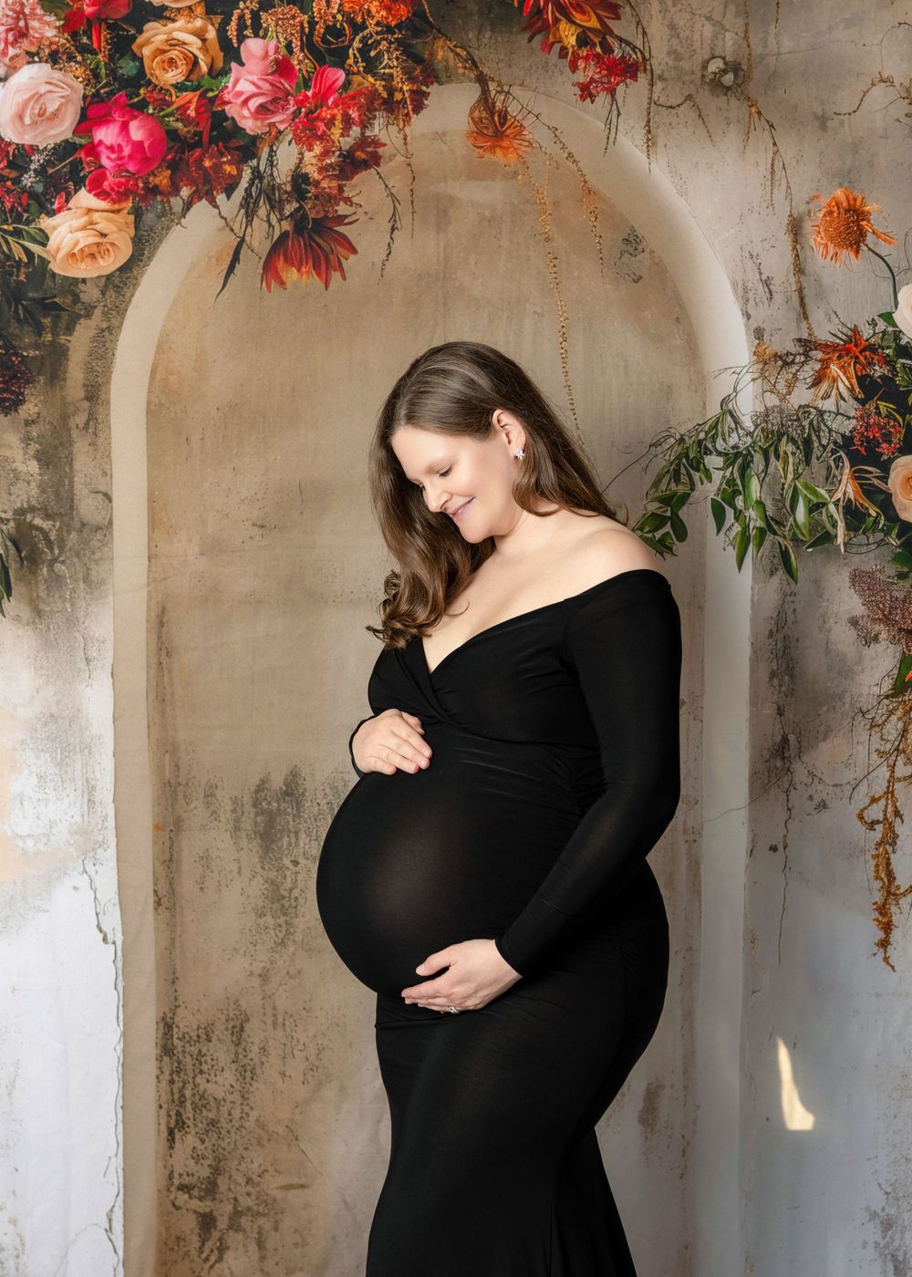 Pregnant woman in black off-shoulder gown cradling belly, posed before floral arch with pink and orange blooms.