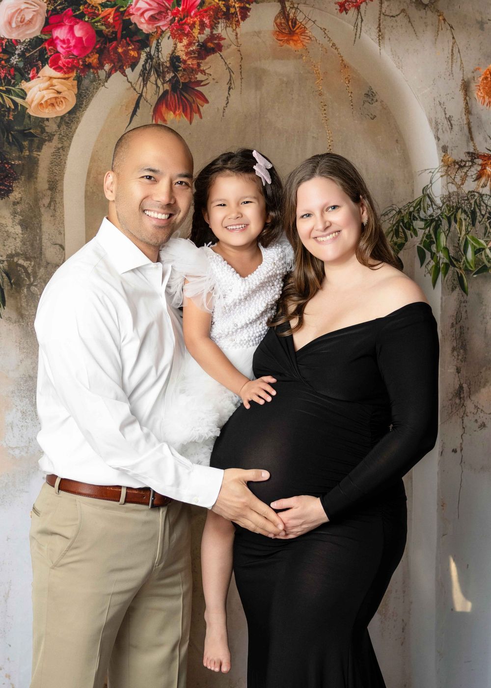 Expecting family maternity portrait with father, young daughter, and pregnant mother in black gown before floral arch backdrop.