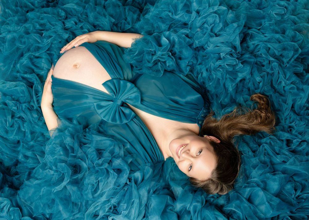 Pregnant woman in teal gown lying on ruffled fabric, cradling baby bump, smiling serenely.